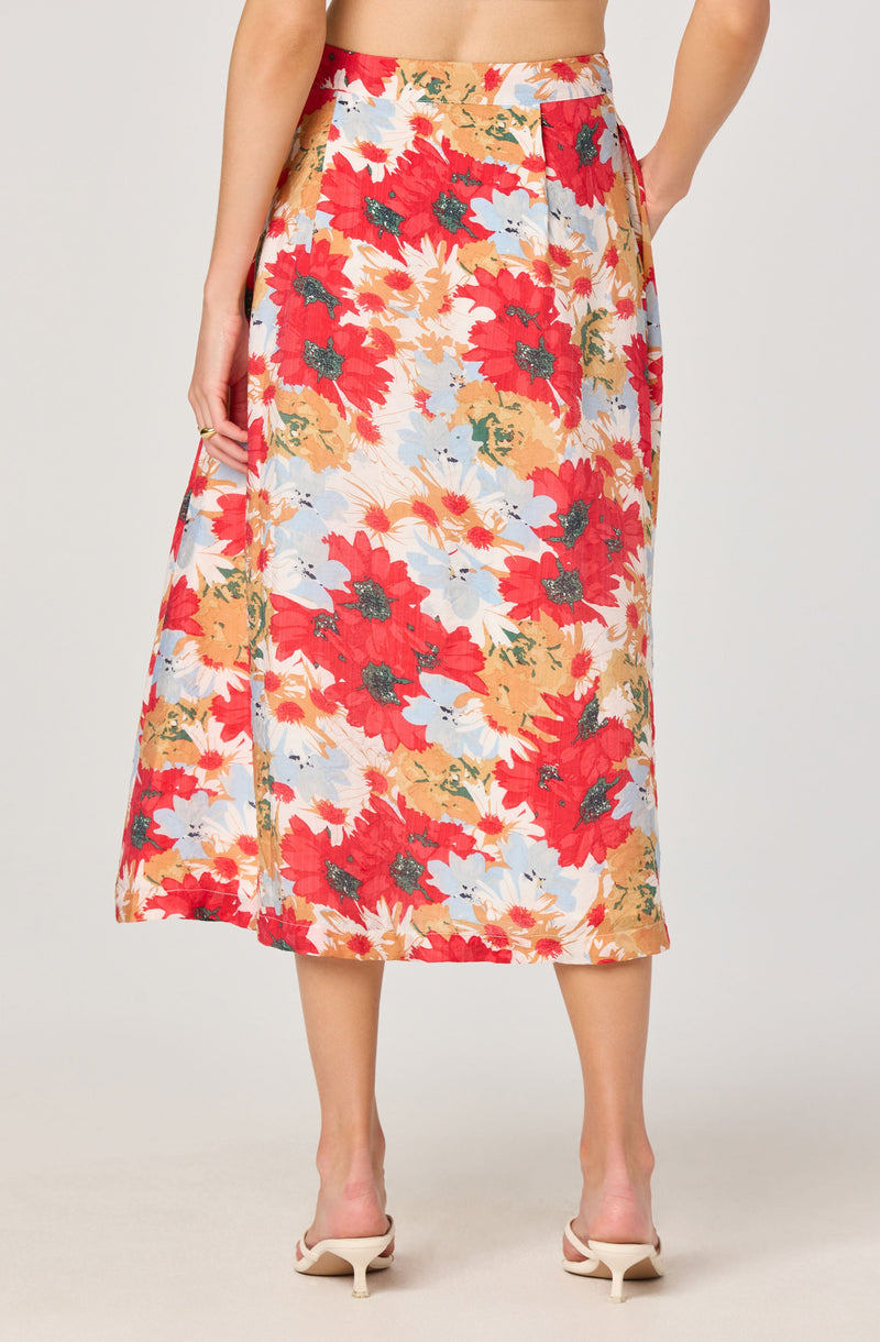 A woman is wearing a long, flowy midi skirt with a vibrant floral pattern. The skirt features large red flowers with dark centers, complemented by smaller white and light blue flowers and green leaves on a light background. She is also wearing beige high-heeled shoes.
