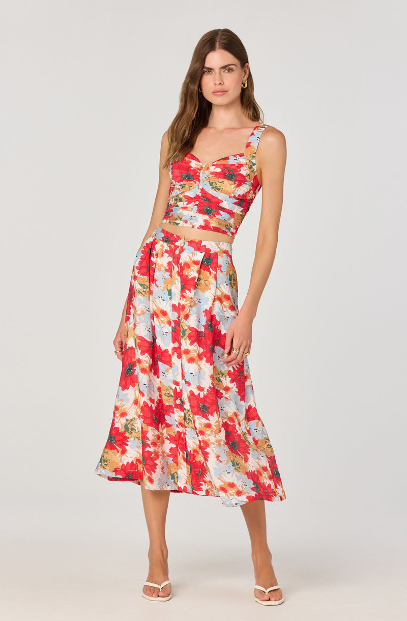 A woman is wearing a sleeveless crop top with a colorful floral pattern featuring shades of red, orange, yellow, blue, and green, with a sweetheart neckline and ruched detailing at the front. She pairs it with a matching high-waisted, A-line midi skirt that also has a vibrant floral print in the same colors, with a button-down front. She completes the outfit with white open-toe heels.