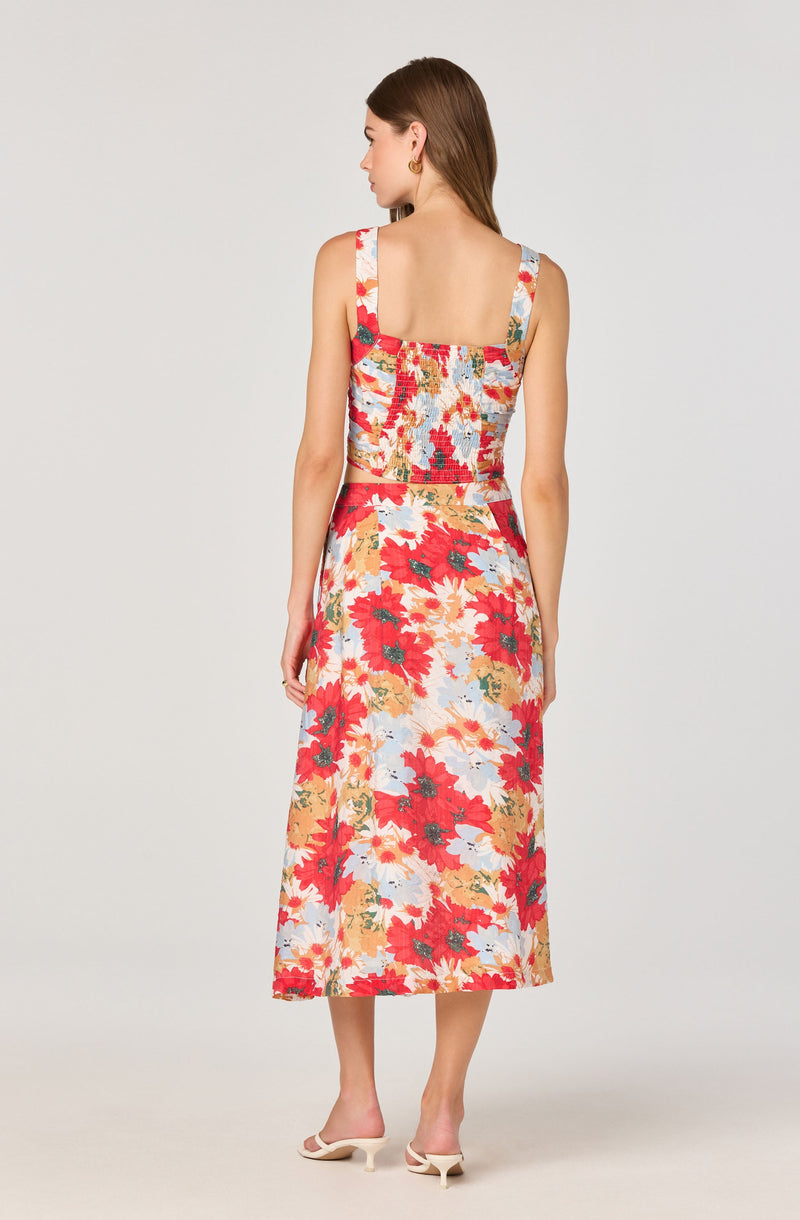A woman is wearing a sleeveless, two-piece floral outfit with a fitted top and a long skirt. The top has wide straps and a straight neckline. The fabric features a colorful floral pattern with dominant red, white, yellow, and green shades. The matching skirt is high-waisted, flowy, and reaches her ankles. She is also wearing white open-toed heels.