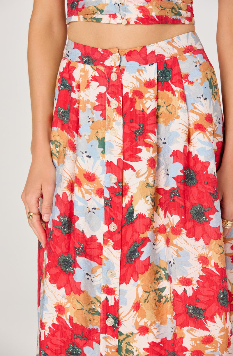 A woman is wearing a matching floral crop top and long skirt set. The top is sleeveless and fitted at the waist, with a colorful pattern of red, orange, light blue, and white flowers. The high-waisted skirt features the same vibrant floral pattern and has a row of white buttons running down the front.
