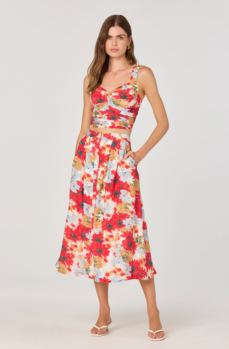 A woman is wearing a matching two-piece set consisting of a sleeveless crop top and a high-waisted, A-line skirt. The fabric features a vibrant floral pattern with large, colorful flowers in shades of red, orange, yellow, and light blue, with green accents. The crop top has a sweetheart neckline and wide shoulder straps, while the skirt has a loose fit and reaches mid-calf length. She is also wearing white high-heeled sandals.