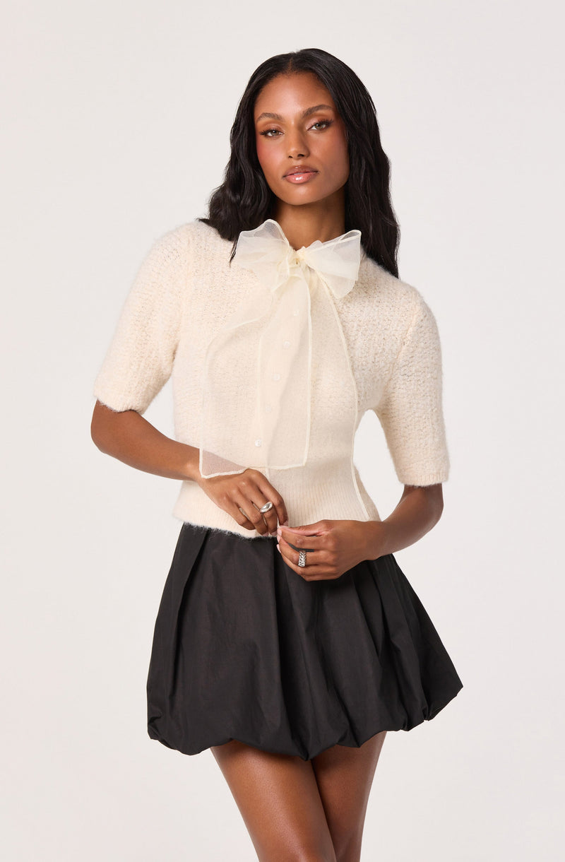 A woman is wearing a short-sleeved, cream-colored knit sweater with a fitted waist. She has a sheer, cream-colored bow tie around her neck, tied in a large bow. She pairs the top with a black, voluminous, bubble-style skirt that sits high on her waist.