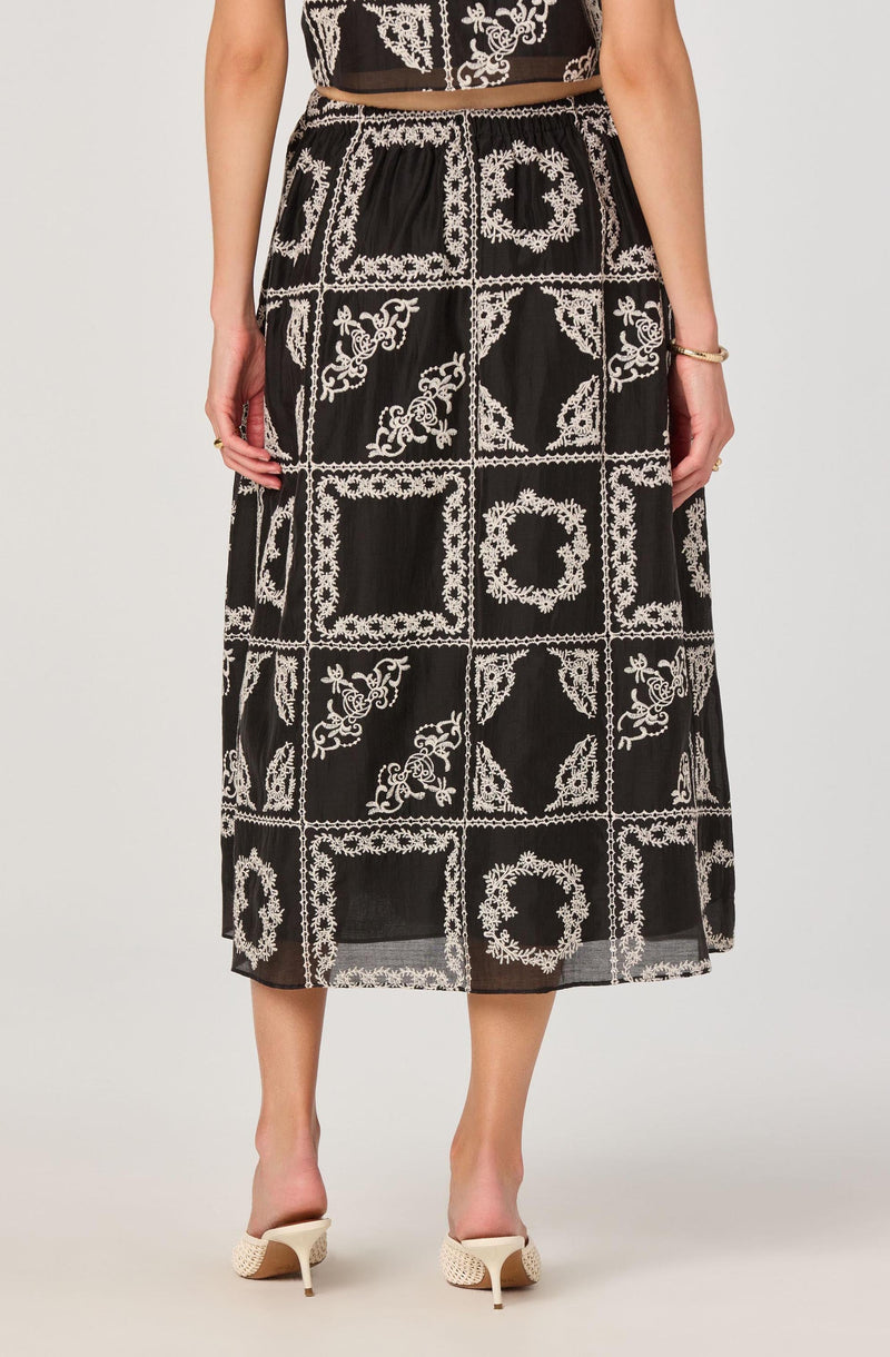 A person is wearing a black midi skirt with a white embroidered geometric and floral pattern, featuring squares and circular motifs. The embroidery design is detailed and covers the entire length of the skirt. The person has gold accessories, including a bracelet and rings, and is wearing white high-heeled shoes.
