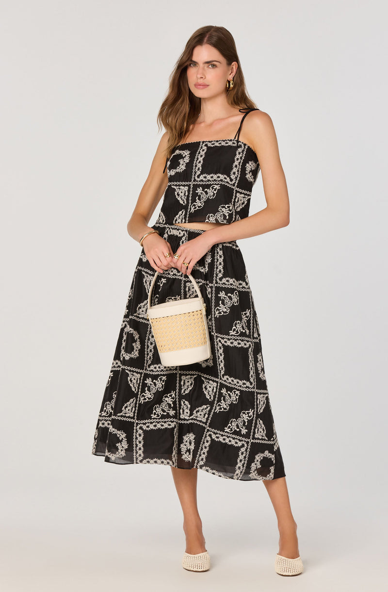 A woman is wearing a black sleeveless jumpsuit with thin spaghetti straps. The jumpsuit features a white floral and geometric pattern throughout. She is holding a beige woven handbag with a white bottom and a rounded handle. She is accessorized with gold hoop earrings and a gold bracelet. She is wearing white woven high-heeled shoes.