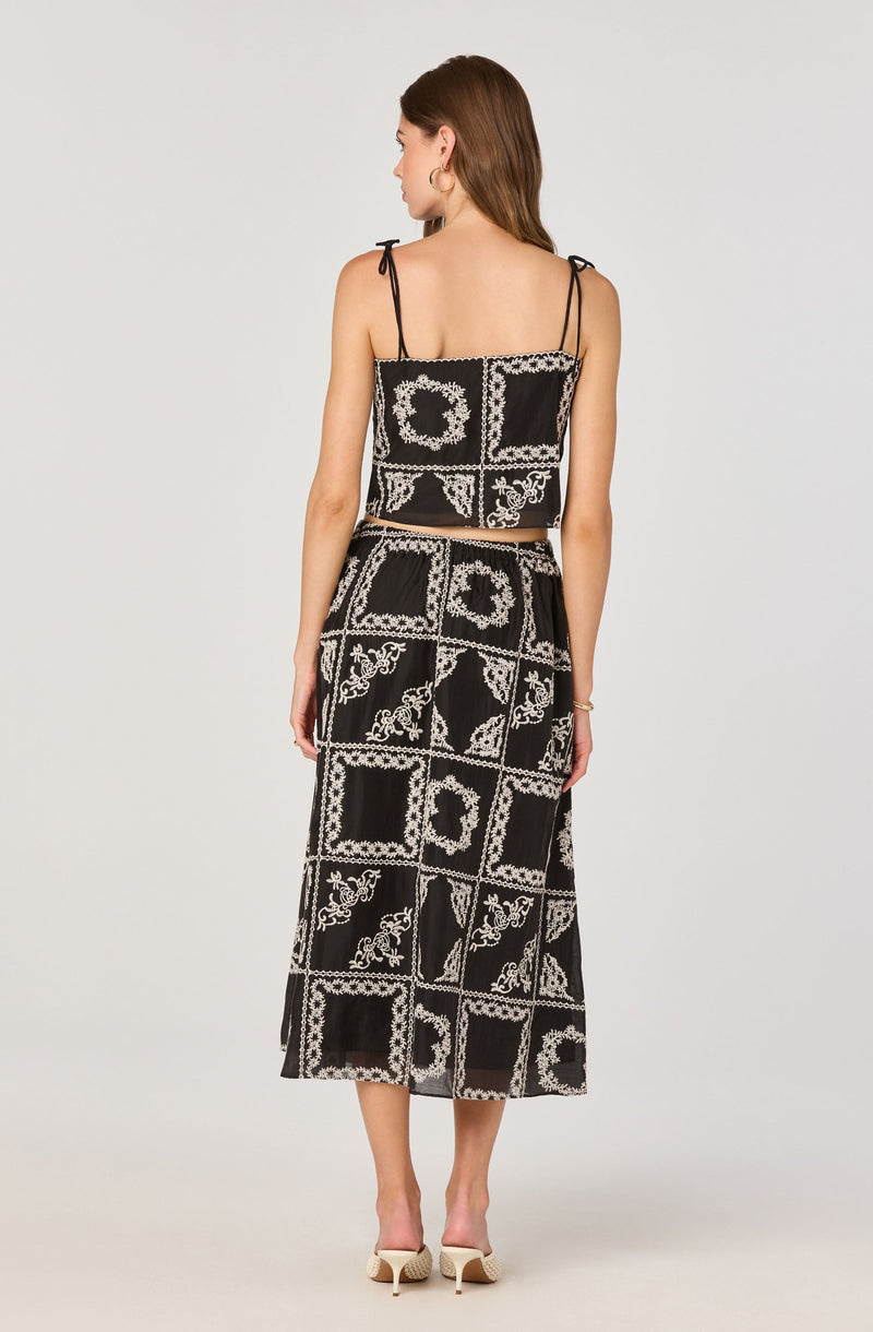 A woman is wearing a two-piece outfit consisting of a sleeveless crop top and a long skirt. The top has thin, adjustable shoulder straps and features a black and white pattern with decorative border designs. The matching high-waisted skirt has a similar pattern, extending to mid-calf length, with a slight slit on one side. She is also wearing nude high-heeled shoes.