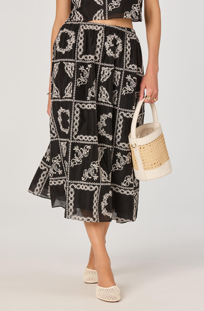 A woman is wearing a black dress with a white embroidered pattern that resembles floral and geometric motifs. The dress has a midi length and a flowy, sheer fabric. She pairs it with beige woven shoes and carries a cream-colored handbag with a woven design on the front. She accessorizes with rings on her fingers.