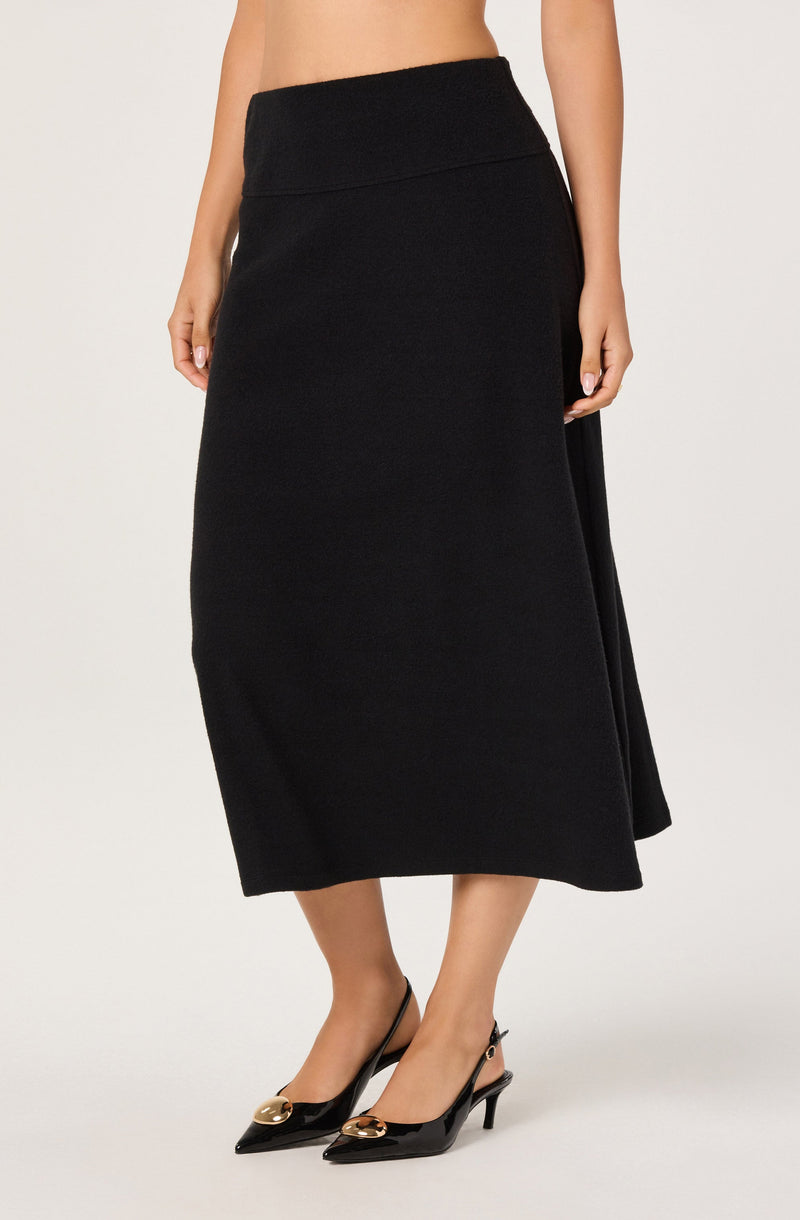 A woman is wearing a long, high-waisted black skirt that extends to mid-calf length. The skirt appears to be made of a textured fabric and has a fitted waistband. She is also wearing black pointed-toe high heels with gold embellishments on the front straps.