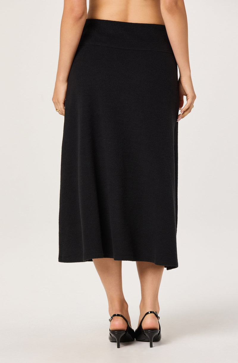 A woman is wearing a high-waisted, ankle-length black skirt with a simple, smooth appearance. She is also wearing black high-heeled shoes with ankle straps.