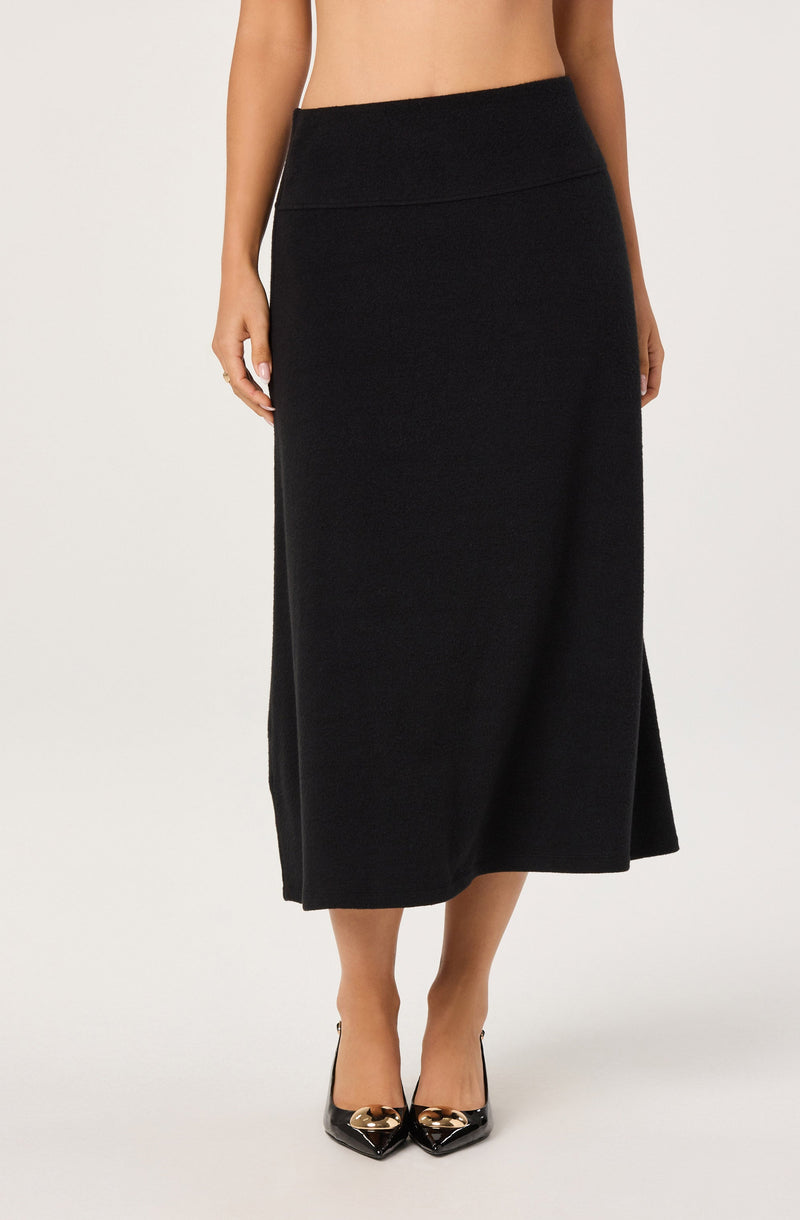 A woman is wearing a high-waisted, ankle-length black skirt with a smooth, slightly fitted silhouette. She is also wearing black pointed-toe high heels with metallic gold accents on the toes.