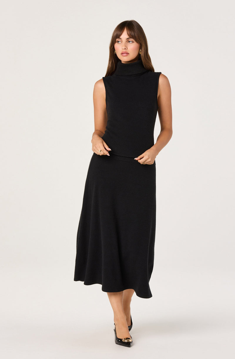 A woman is wearing a sleeveless, black turtleneck top made of a ribbed knit material. She pairs it with a black, ankle-length, A-line skirt that drapes softly down to her ankles. She is also wearing black pointed-toe heels with a gold accent on the toe box.
