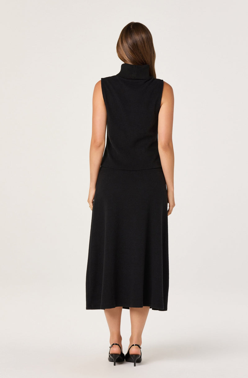 A woman is wearing a sleeveless black turtleneck top and a matching black midi-length skirt. She has long brown hair and is standing with her back to the camera. She is also wearing black high-heeled shoes.