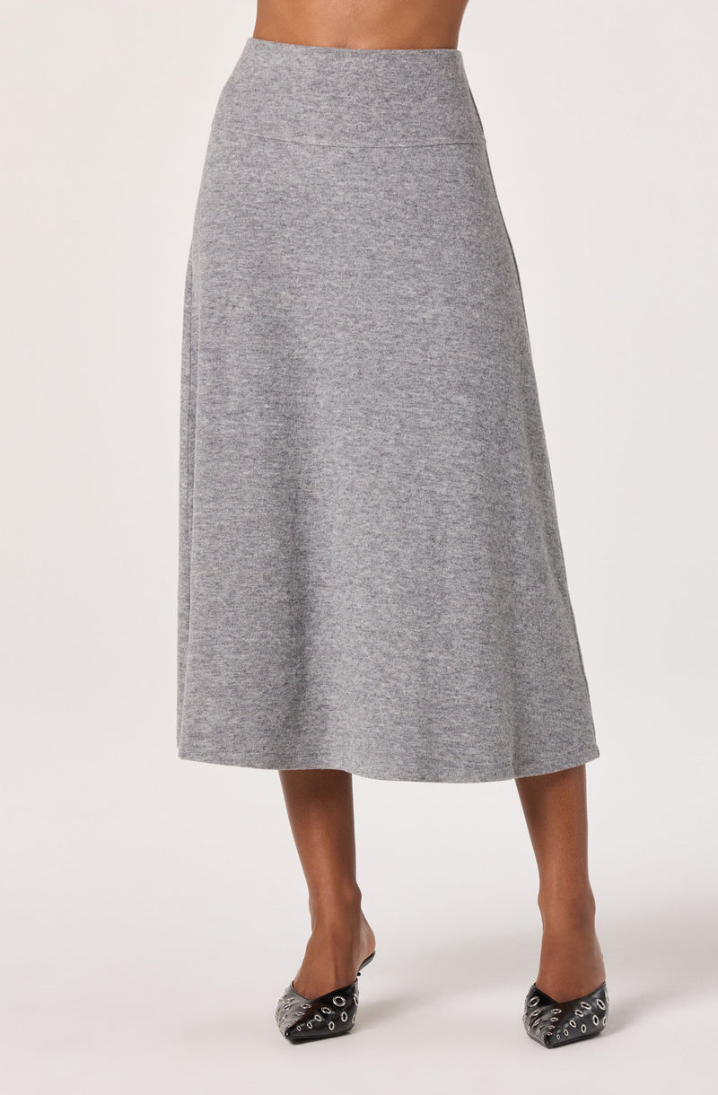 A woman is wearing a high-waisted, ankle-length, heather gray skirt made of a soft, knit fabric. She is also wearing black pointed-toe shoes with silver eyelet details.