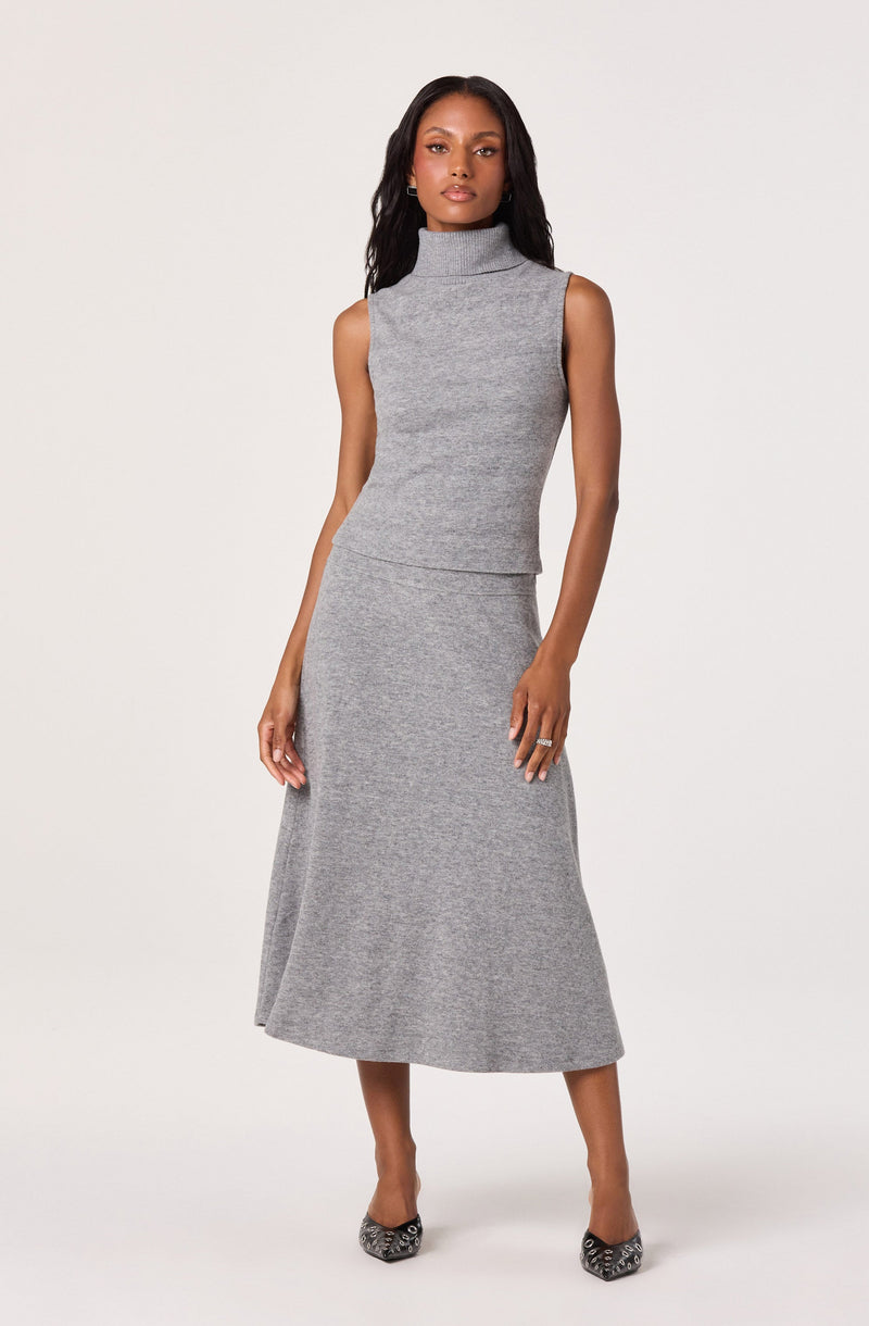 A woman is wearing a sleeveless, high-neck, knit top and a matching knit midi skirt, both in a light gray color. She pairs the outfit with pointed-toe shoes that have a patterned design.