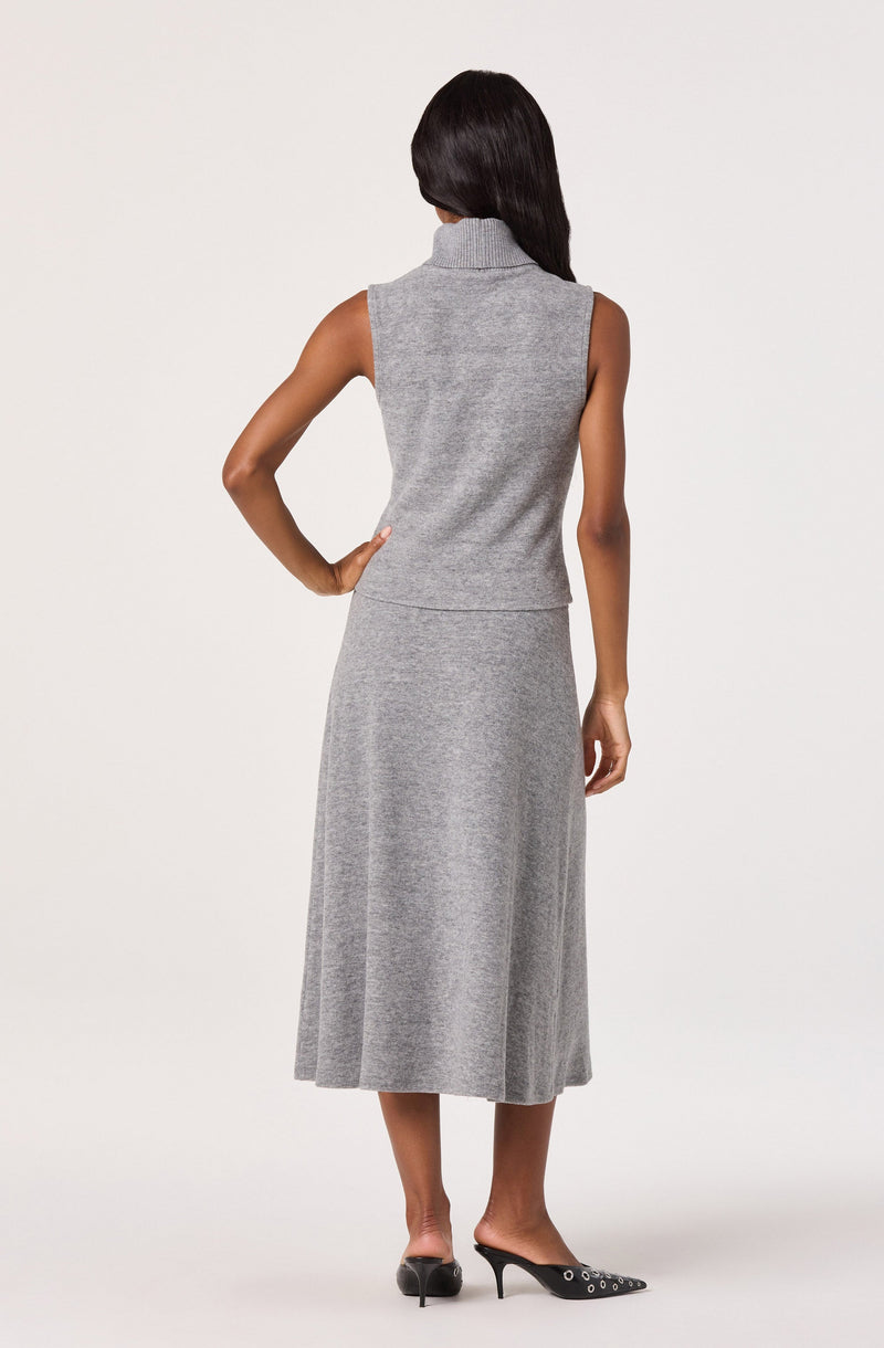 A woman is wearing a sleeveless, light gray knit vest with a high collar, paired with a long, matching gray knit skirt that reaches her mid-calf. She is also wearing black high-heeled shoes with pointed toes and decorative eyelets.