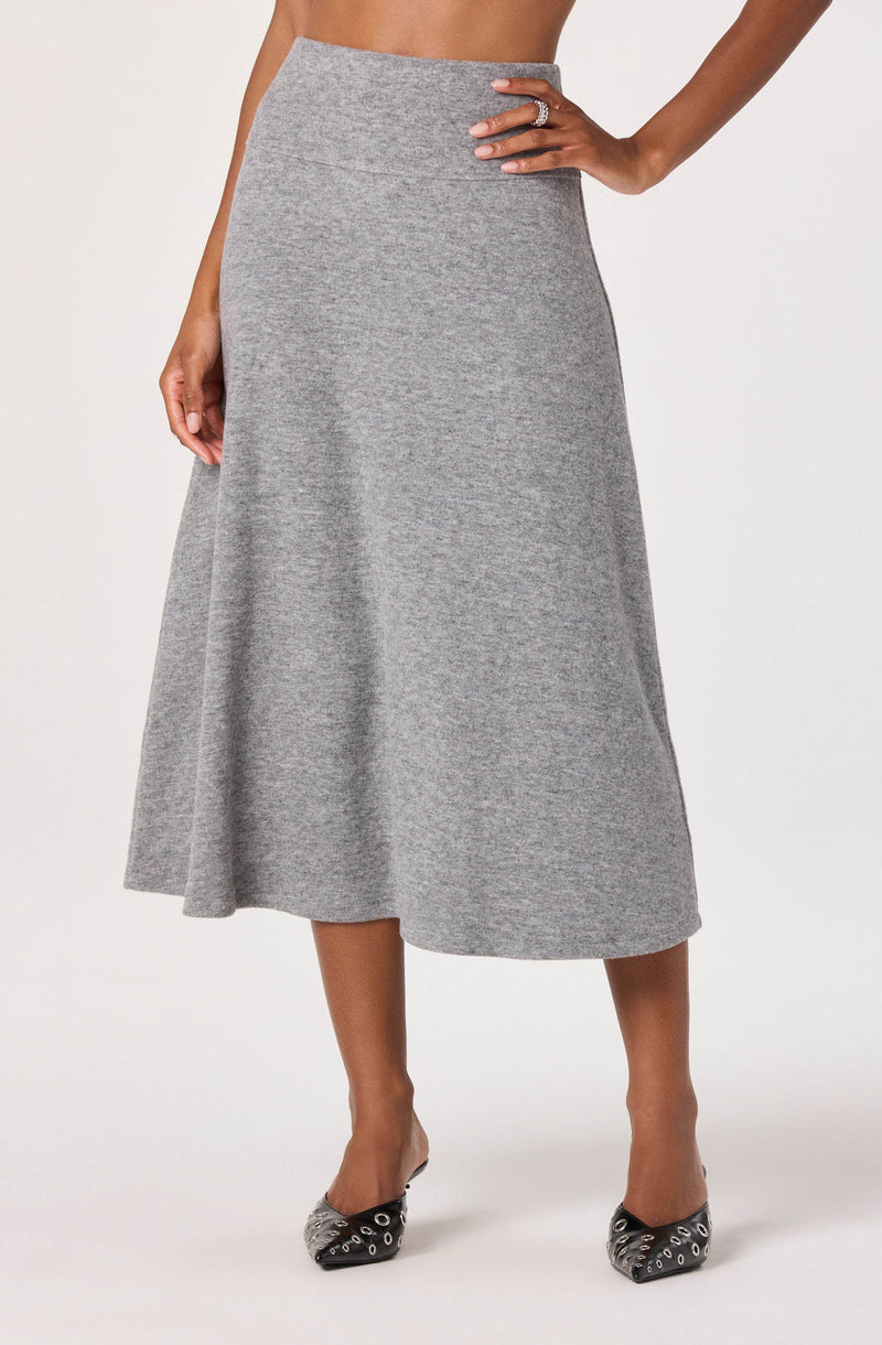 A woman is wearing a high-waisted, A-line, ankle-length gray skirt made of a soft, textured fabric. She has one hand on her hip, showing a ring on her finger. She is also wearing black shoes with silver eyelet details.
