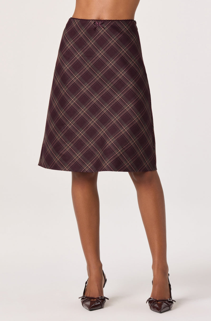 A woman is wearing a knee-length, high-waisted skirt with a tartan plaid pattern in shades of maroon, green, and beige. The skirt has a small, decorative bow at the waistband. She is also wearing dark, glossy high heels with a bow detail on the front.