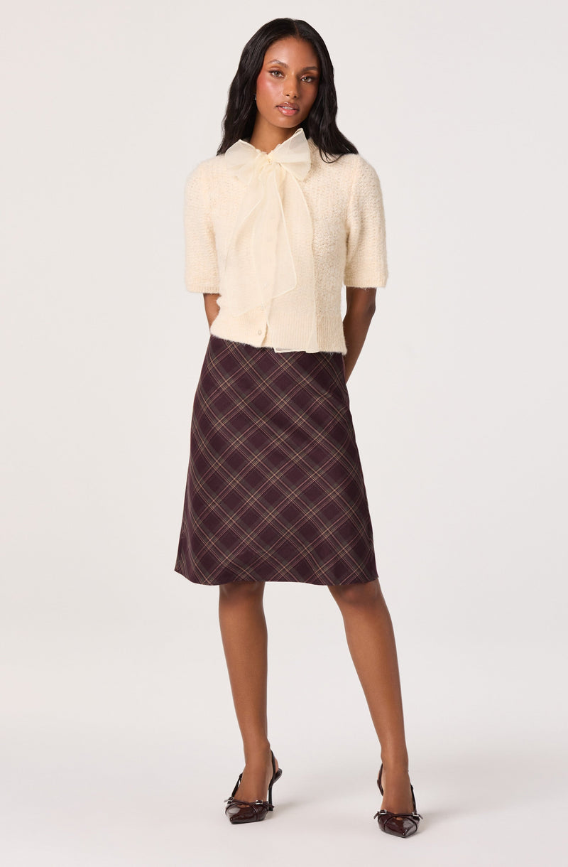A woman is wearing a short, cream-colored, textured knit cardigan with a large bow at the collar. She has paired it with a high-waisted, knee-length A-line skirt featuring a plaid pattern in shades of burgundy, yellow, and green. She completes the look with pointed-toe heels with a bow detail.