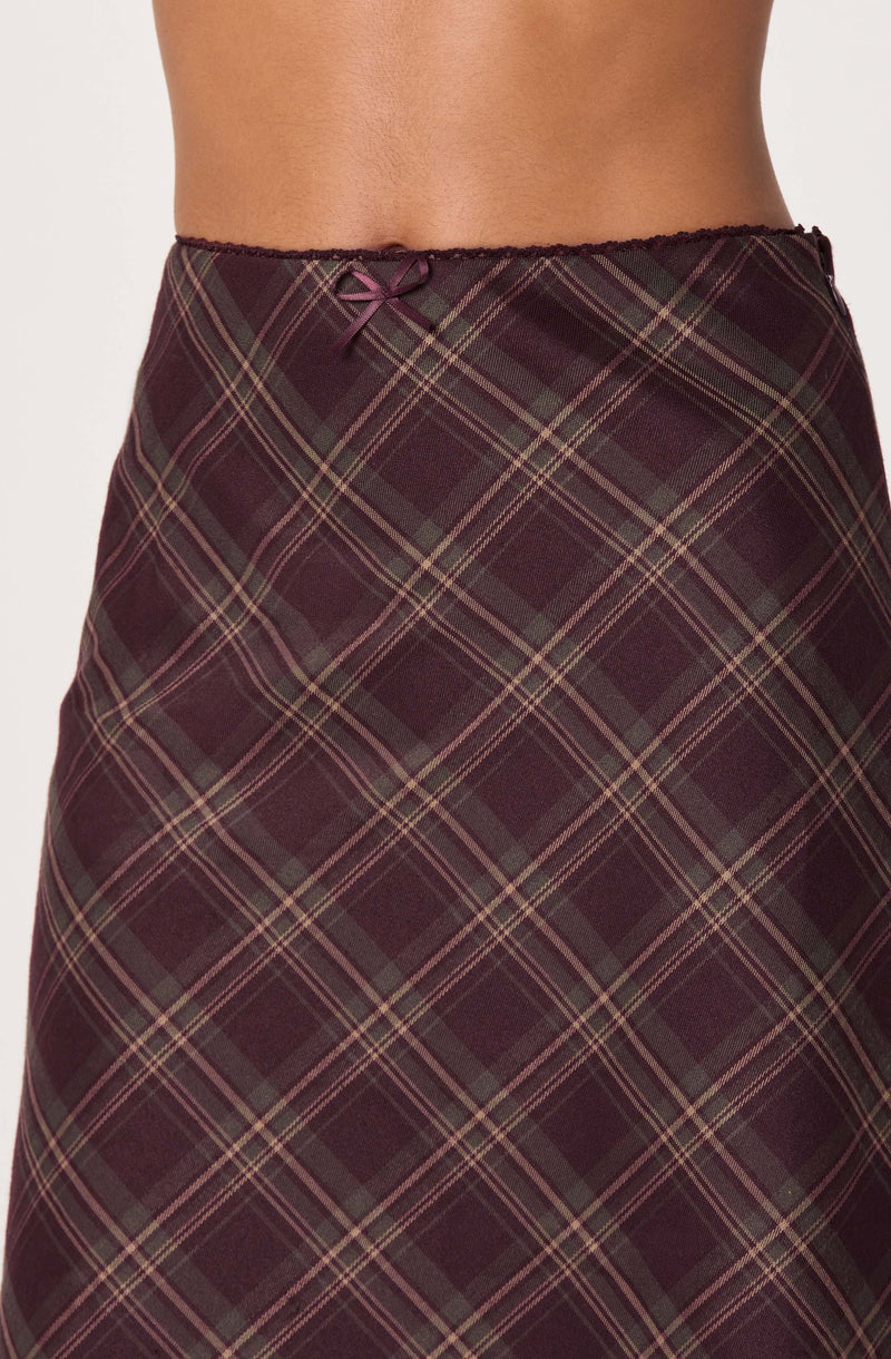 A close-up of a person wearing a plaid skirt with a dark maroon and green pattern, featuring thin yellow and red lines. The waistband is elastic with a small decorative bow at the center.