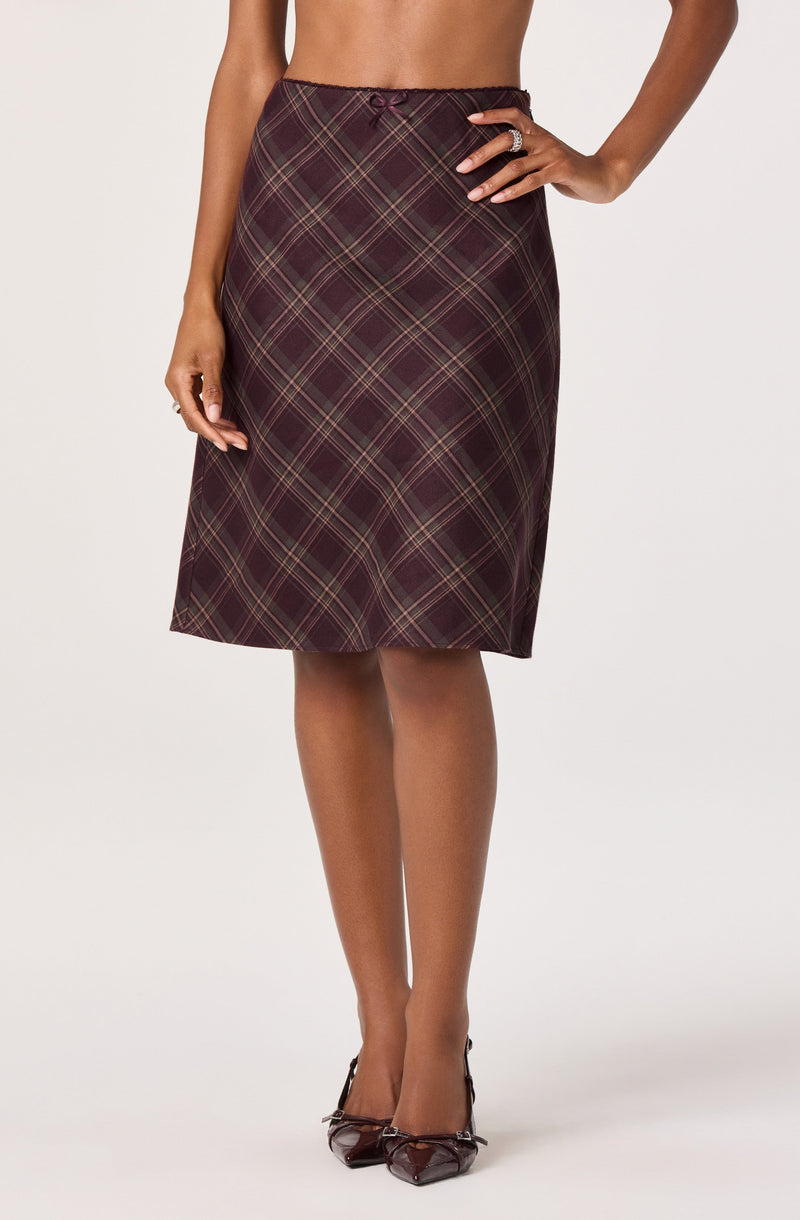 A woman is wearing a knee-length, high-waisted skirt with a plaid pattern in shades of maroon, beige, and black. She has one hand on her hip and is wearing a large ring on her finger. She is also wearing a pair of dark-colored, pointed-toe heels.