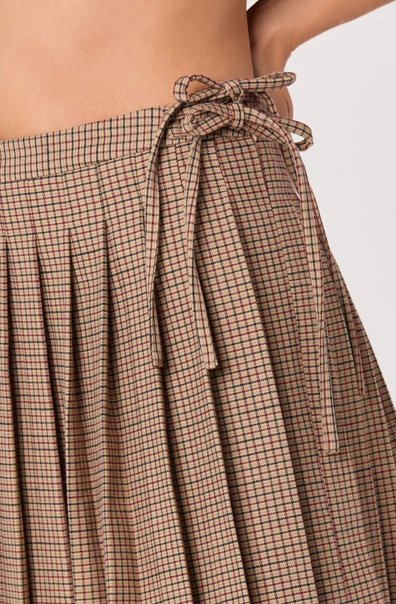 A close-up of a pair of pleated, high-waisted pants with a small checkered pattern in shades of beige, red, and black. The waistband has a drawstring tie made from the same fabric, tied in a bow at the front.