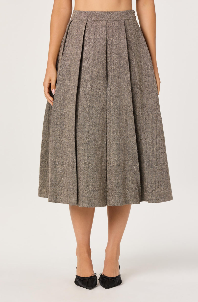 A woman is wearing a high-waisted, full-length brown skirt with a subtle checkered pattern. The skirt features wide pleats that run vertically from the waistband to the hem, creating a voluminous silhouette. She pairs the skirt with black pointed-toe shoes that have bow details on the front.