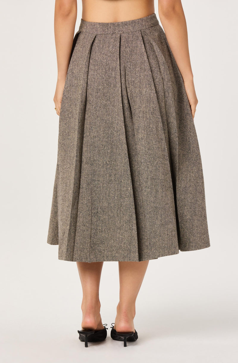 A woman is wearing a high-waisted, ankle-length skirt made of a textured, brownish plaid fabric with pleats running down the front. She is also wearing black open-toed heels with ankle straps.