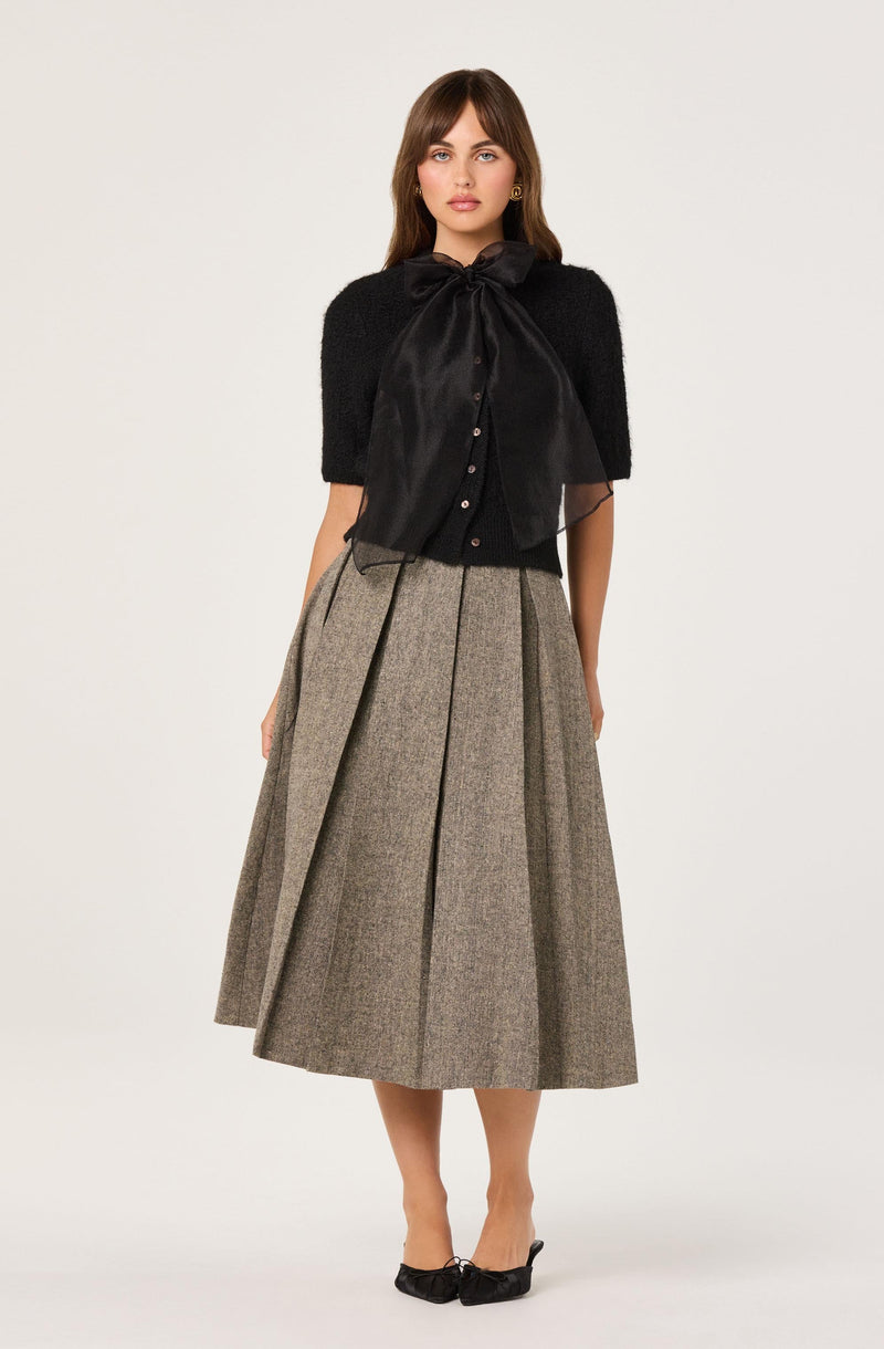 A woman is wearing a black short-sleeved top with a large satin bow at the neckline and small buttons down the front. She pairs it with a high-waisted, pleated, midi-length skirt made of a beige and brown checkered fabric. She is also wearing black pointed-toe heels with a bow detail.