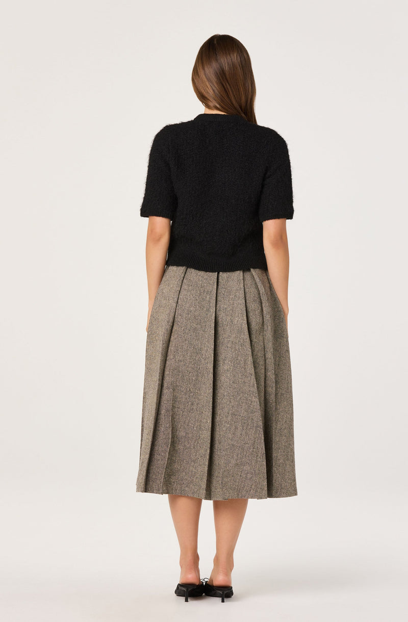 A woman is wearing a black, short-sleeved, fuzzy sweater and a high-waisted, ankle-length, pleated skirt made of plaid fabric in shades of gray and brown. She is also wearing black heeled shoes.