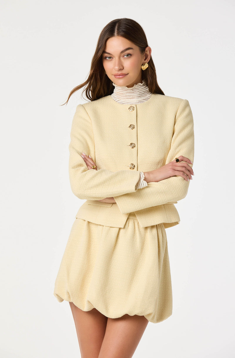 A woman wearing a light cream-colored textured suit consisting of a fitted, long-sleeved jacket with gold buttons and a matching skirt with a puffy, gathered hem. She has on a ruffled, high-neck blouse underneath the jacket, large gold earrings, and a ring on her right hand.