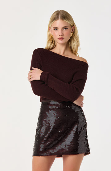A young woman is wearing a maroon, off-the-shoulder, knit sweater with long sleeves. She pairs it with a high-waisted, dark sequined mini skirt that has a shiny, reflective surface.