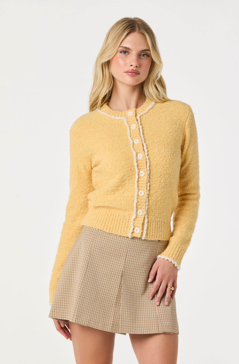 A woman is wearing a yellow, textured, long-sleeve cardigan with a round neckline and white piping detail along the edges and buttons. She pairs it with a beige plaid mini skirt.