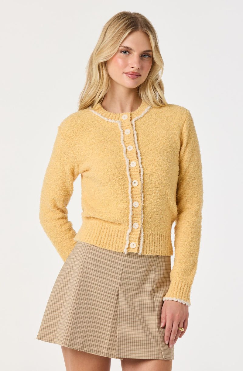 A woman is wearing a yellow, textured knit cardigan with white scalloped trim along the neckline, front buttons, and cuffs. She pairs it with a beige plaid skirt.