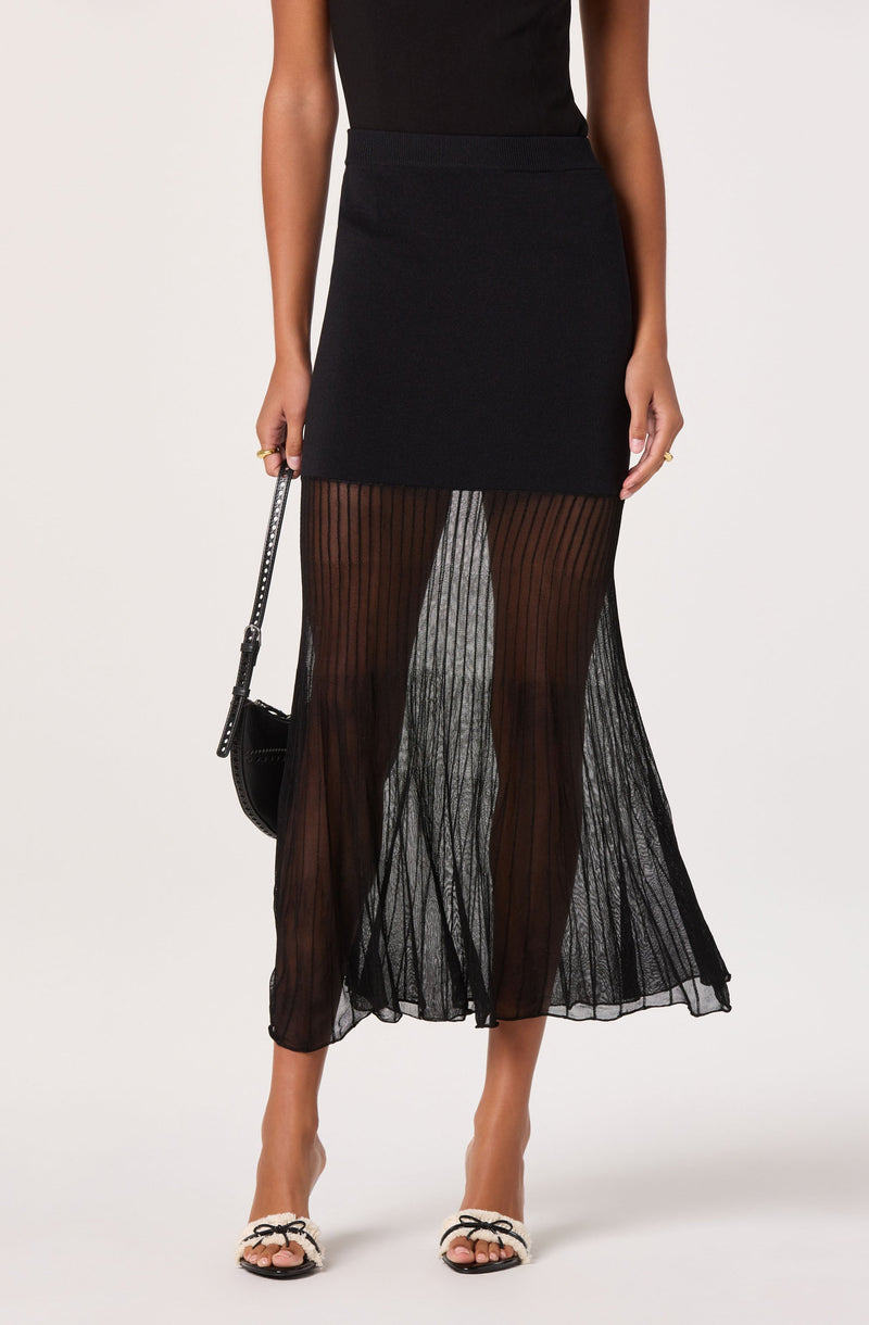 A person wearing a black sleeveless top tucked into a black high-waisted skirt that is short and fitted at the top. They are also wearing sheer, black, vertically striped wide-leg pants that extend to the floor. The person is holding a small black handbag with a chain strap and is wearing open-toed sandals with a bow detail on the front.