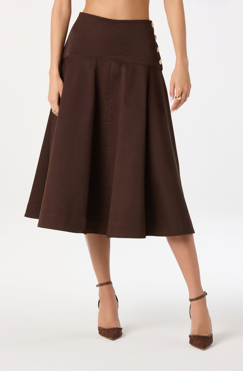 A woman is wearing a high-waisted, dark brown midi skirt with a wide waistband. The skirt features a row of decorative buttons on the side of the waistband and has a full, A-line silhouette with pleats, creating a voluminous look. She is also wearing brown pointed-toe heels with ankle straps.