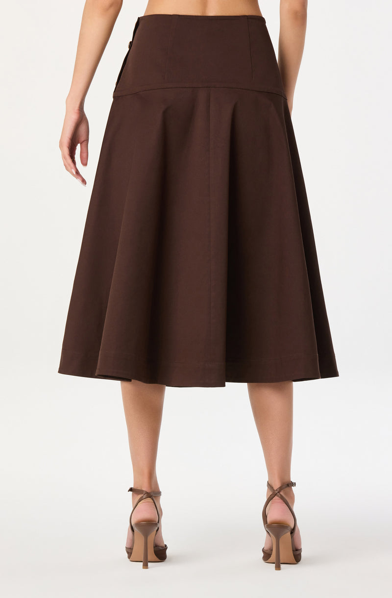 A woman is wearing a high-waisted, dark brown, A-line midi skirt with a wide waistband and pleats that add volume and shape. She is also wearing strappy nude high-heeled sandals.