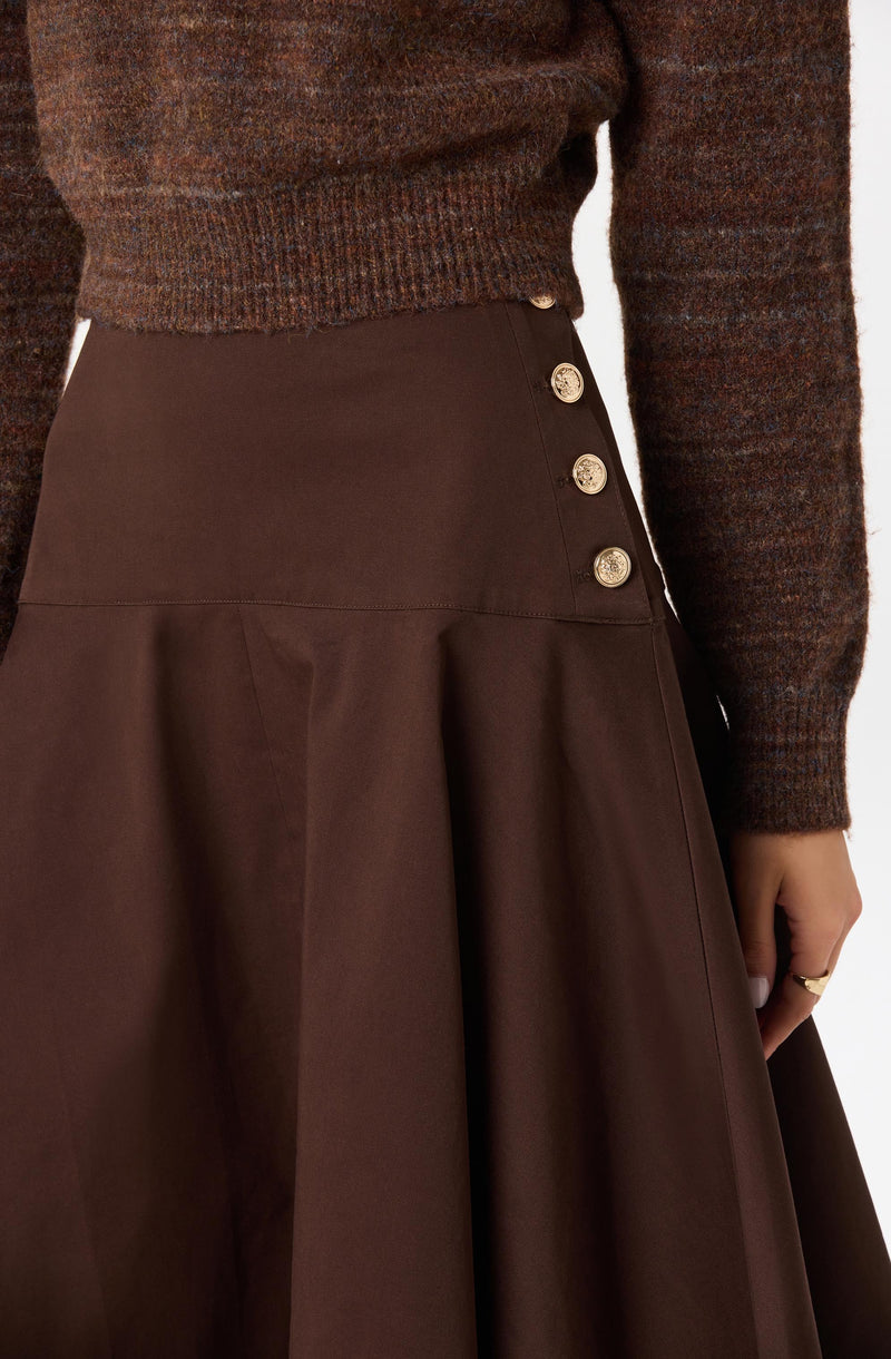 The image shows a person wearing a dark brown, high-waisted skirt with a row of four decorative gold buttons on the side. The skirt has a pleated design below the buttons. The person is also wearing a multicolored, textured sweater with shades of brown, red, and green, with the sweater tucked into the skirt. The person's hand, with a ring on the finger, is resting by their side.