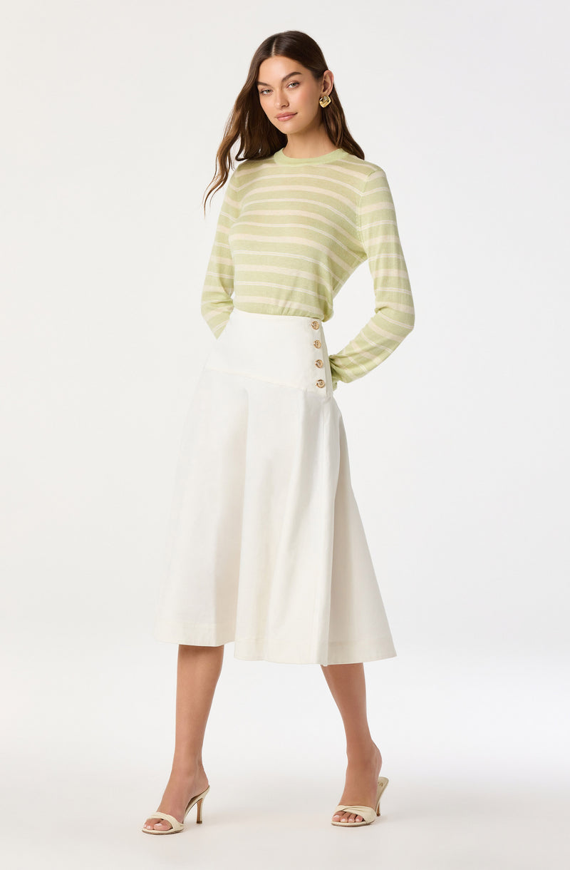 A woman is wearing a light green and white striped long-sleeve top with a round neckline. She pairs it with a high-waisted, white mid-length skirt featuring side buttons. She completes the outfit with beige open-toe high heels.