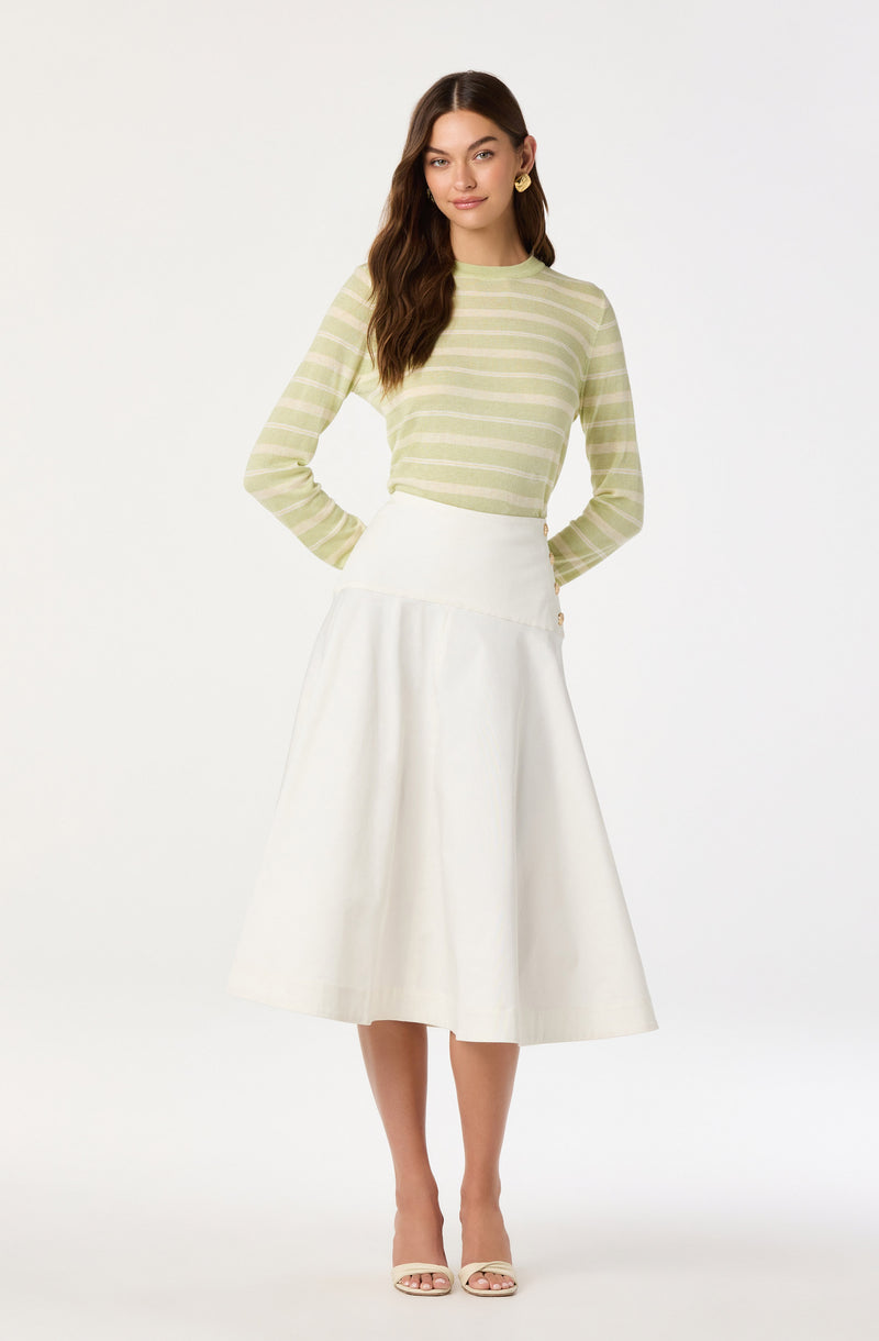 A woman is wearing a light green and cream horizontal striped long-sleeve top with a round neckline. She pairs it with a high-waisted, white, midi-length A-line skirt that reaches below her knees. She completes the outfit with a pair of nude open-toe heels.