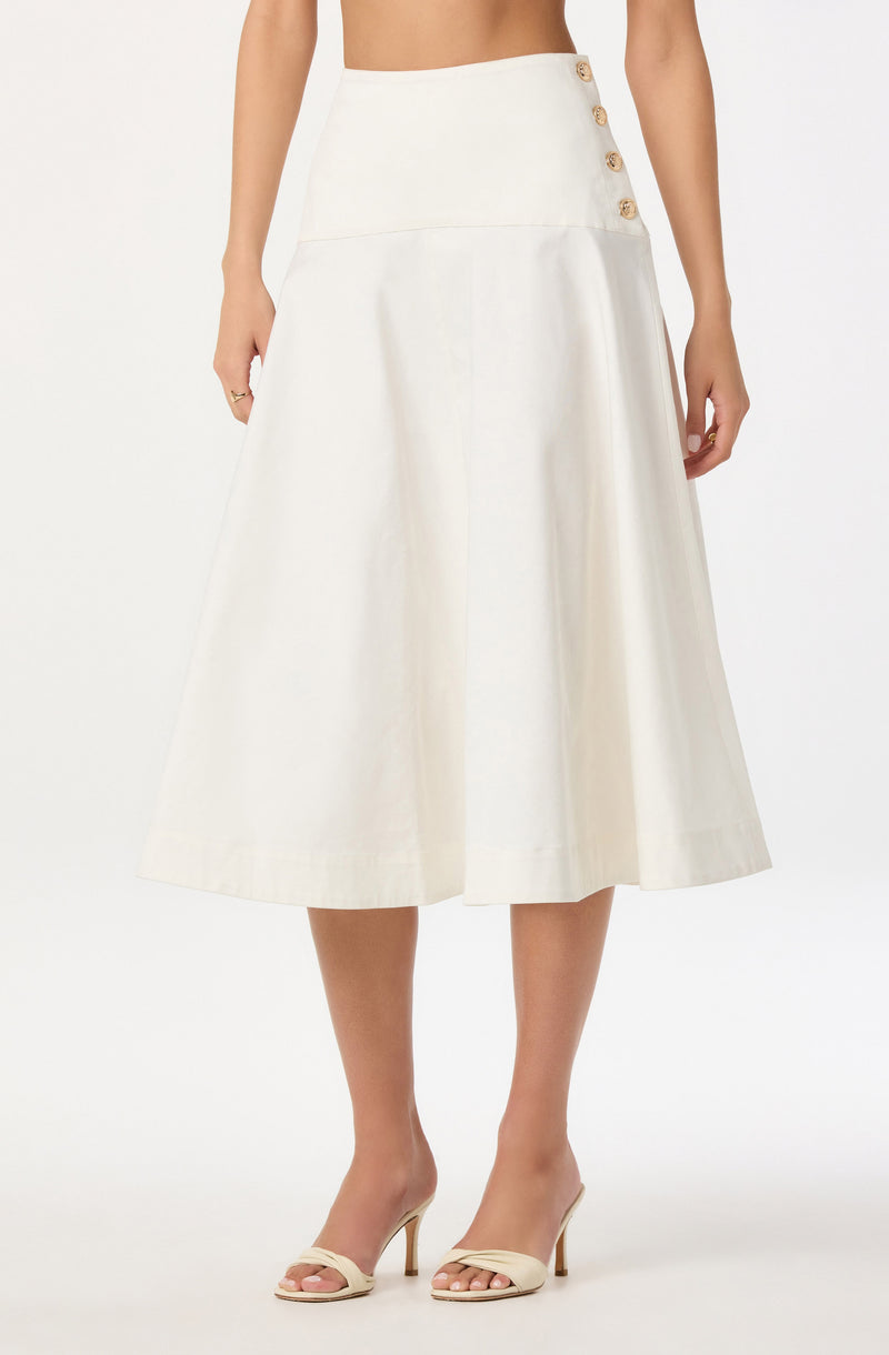A woman is wearing a high-waisted, cream-colored skirt with a wide waistband featuring a row of four decorative buttons on the right side. The skirt is midi-length, flaring out slightly from the waist to below the knees. She is also wearing open-toe beige high-heeled sandals.