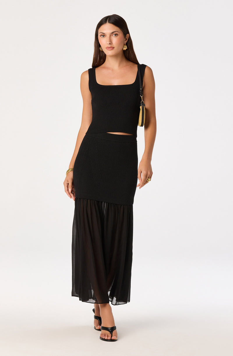 Woman in a black sleeveless ribbed crop top and a matching black sheer maxi skirt.