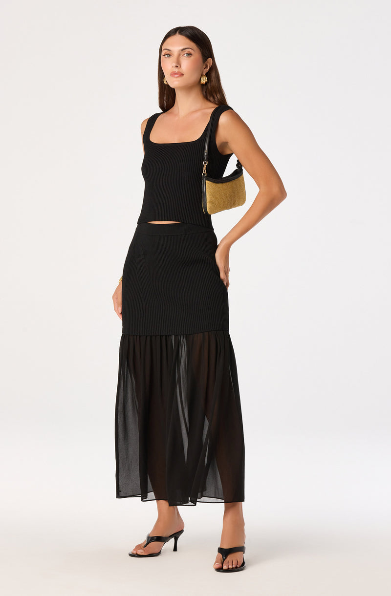 Woman wearing a black sleeveless ribbed top and a black sheer skirt with a ruffled hem, carrying a small two-tone shoulder bag.