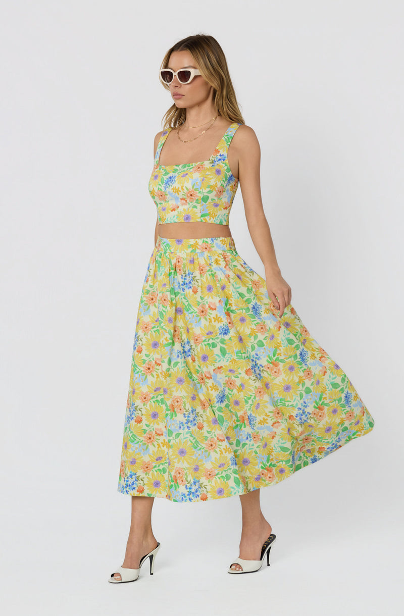 Woman wearing a colorful floral two-piece outfit with a crop top and long skirt, paired with white high heels and sunglasses.