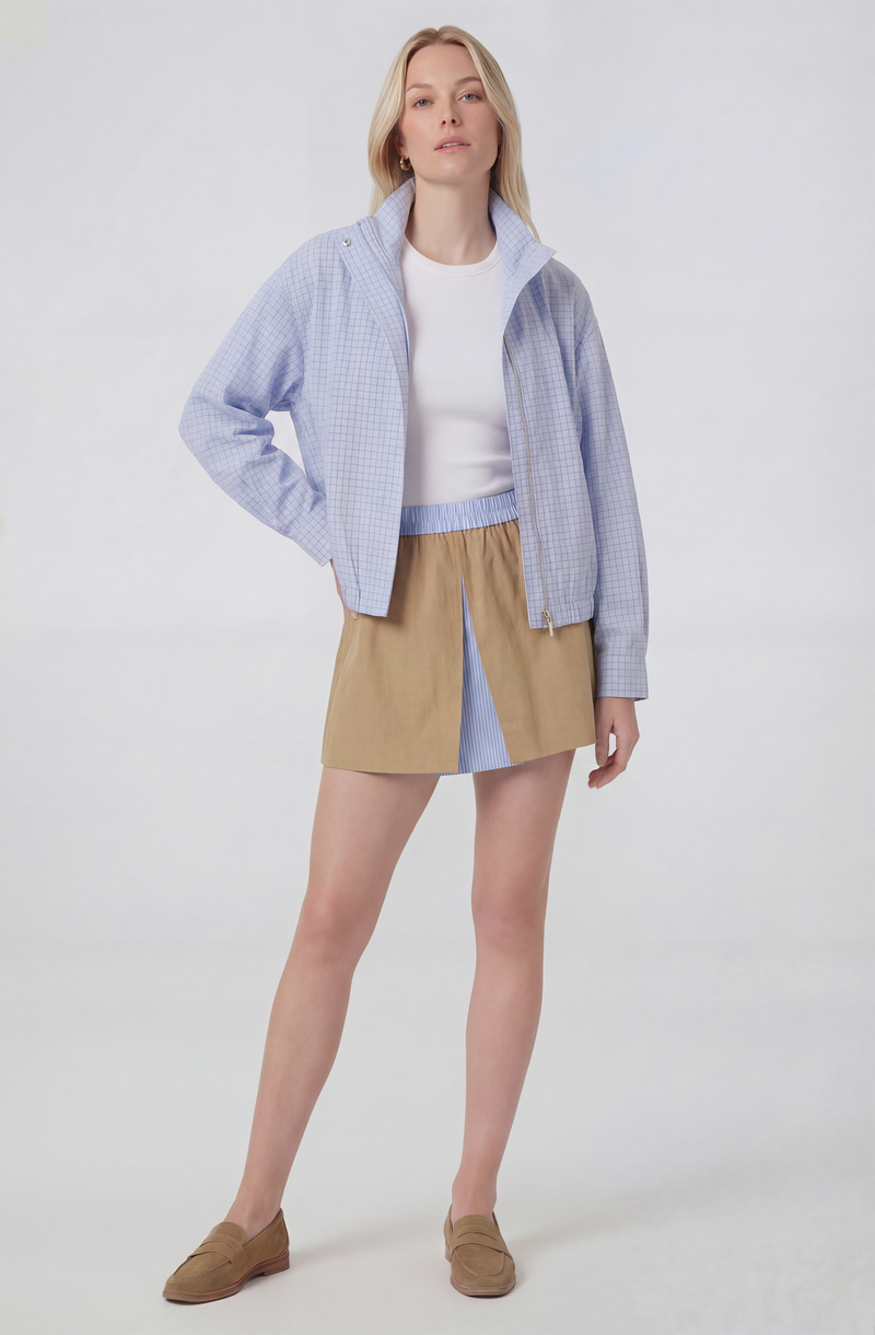 Woman wearing a light blue checkered jacket, white t-shirt, beige and blue skirt, and tan shoes.