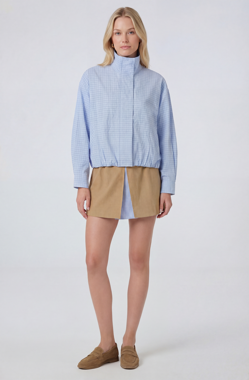 Woman wearing a light blue checkered zip-up jacket with a high collar and tan skirt with a blue striped panel.