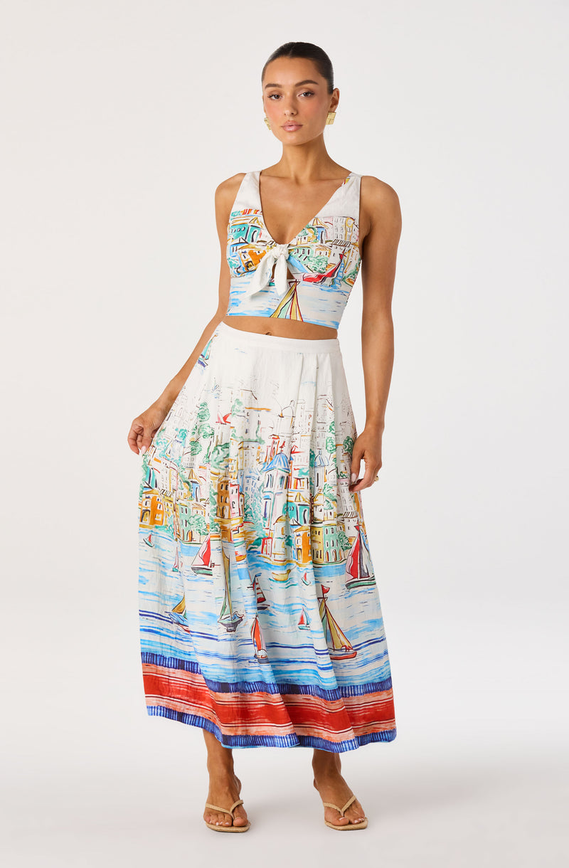 Woman wearing a sleeveless, colorful print crop top with a front tie and a matching long skirt with a seaside town and sailboats pattern.
