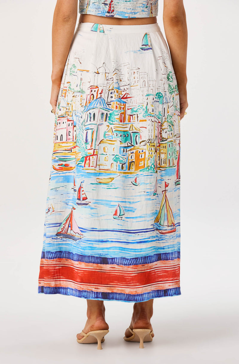 White maxi skirt with colorful cityscape and sailboat print, paired with beige heels.