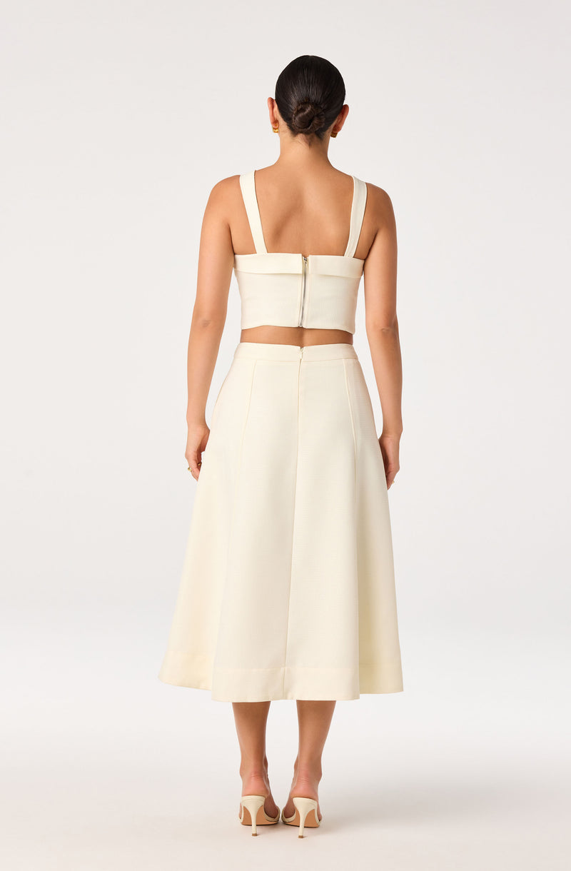 Woman wearing a cream-colored crop top with wide straps and a matching high-waisted, midi skirt.
