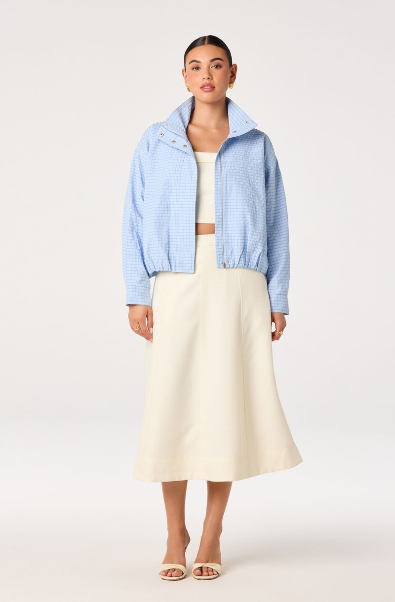 Woman in light blue checkered jacket, cream top, and cream midi skirt, standing against a white background.