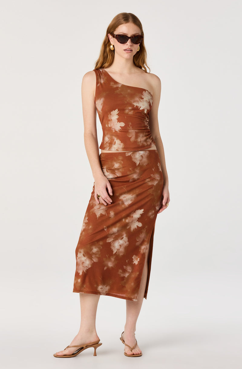 Woman in a brown tie-dye one-shoulder top and matching midi skirt with a side slit, wearing sunglasses and heels.