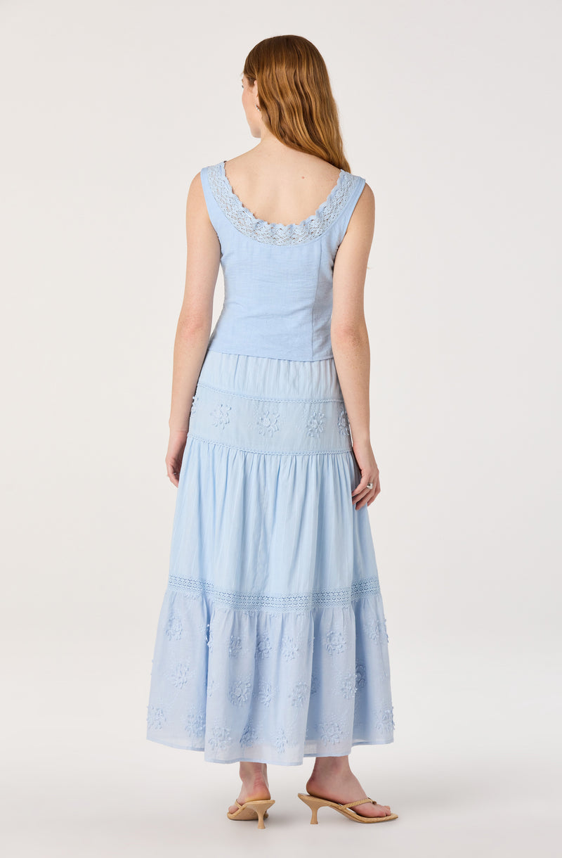 Woman in a light blue sleeveless dress with embroidered lace details, tiered skirt, and beige open-toe heels.